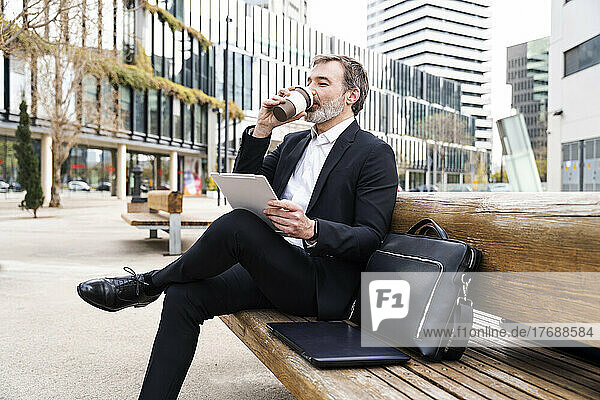 Businessman drinking coffee sitting with tablet PC on bench outside office building