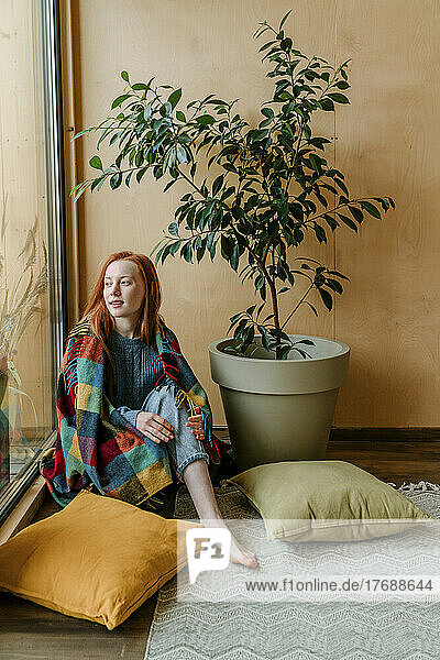 Young woman wrapped in blanket looking through window
