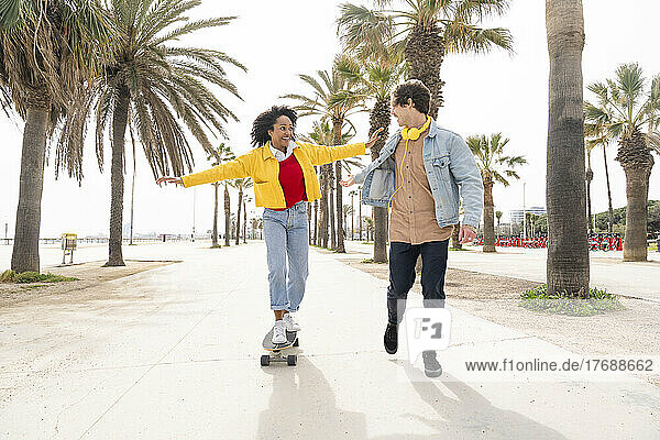 Glücklicher Mann und Frau beim Skateboarden vor Palmen auf dem Fußweg