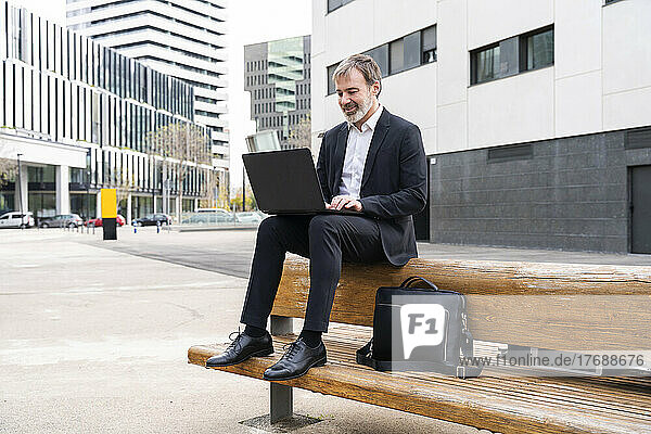Smiling businessman using laptop sitting on bench in front of office building