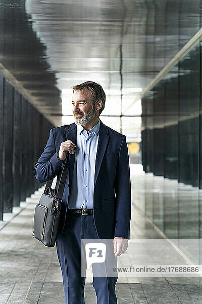 Smiling mature businessman with laptop bag standing in corridor