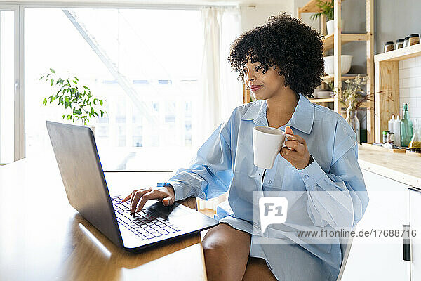 Smiling woman holding coffee cup using laptop sitting at kitchen island