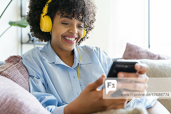 Happy woman wearing headphones using smart phone on sofa at home