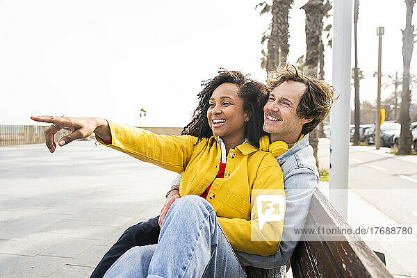 Happy woman sitting on wooden bench with man pointing at footpath