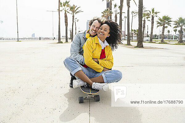 Happy man hugging woman sitting on skateboard in front of trees