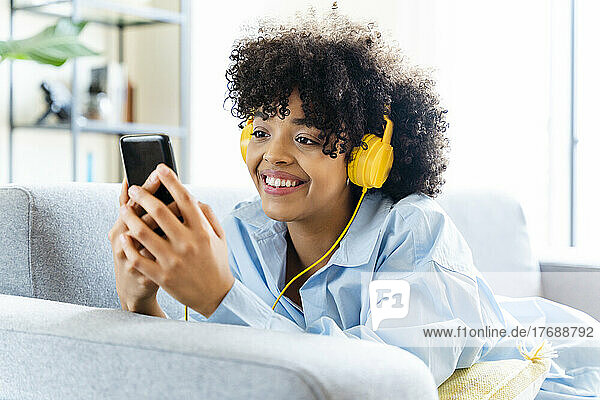 Happy woman with headphones using smart phone lying on sofa at home