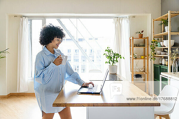 Happy woman with coffee cup using laptop at kitchen island