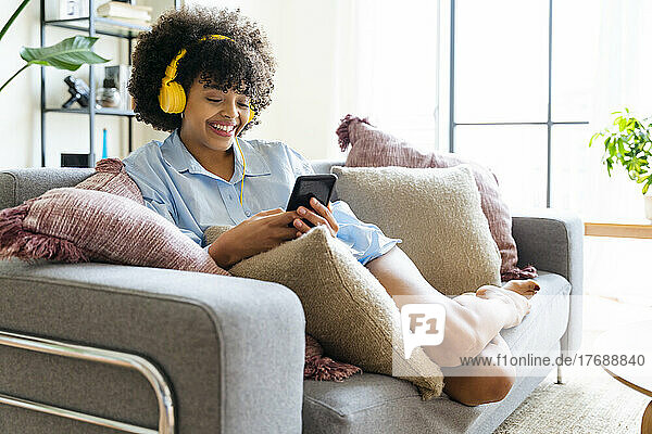 Happy young woman wearing headphones using smart phone on sofa at home
