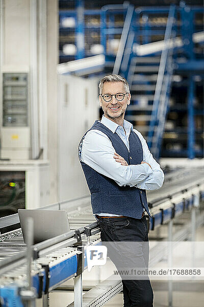 Happy businessman with arms crossed by conveyor belt at factory