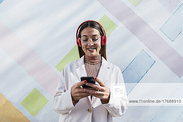 Smiling young woman wearing wireless headphones using mobile phone standing in front of wall