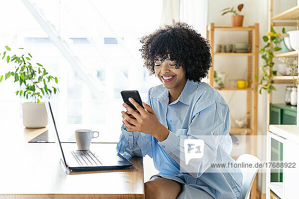 Happy young woman using smart phone sitting at kitchen island