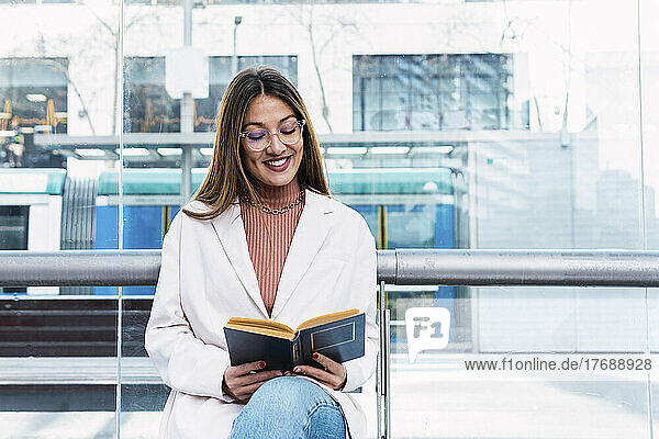 Smiling woman reading book sitting at tram station