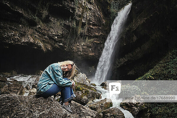Happy mature woman sitting on rock in front of waterfall