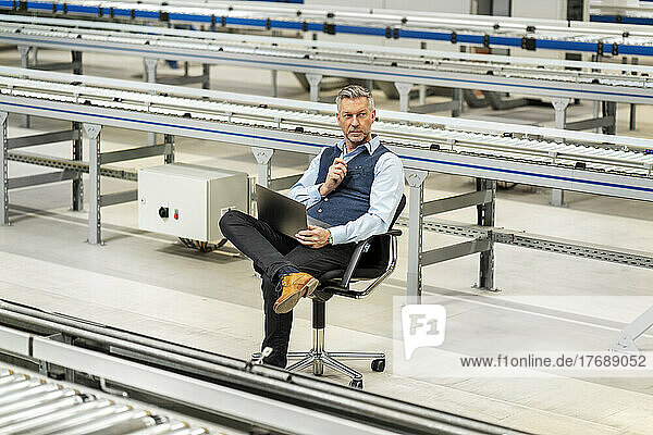 Businessman with laptop sitting amidst conveyor belt at factory
