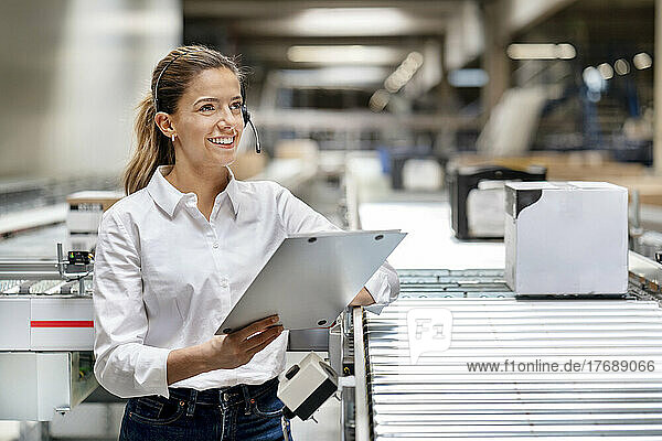 Happy businesswoman with clipboard wearing headset in factory