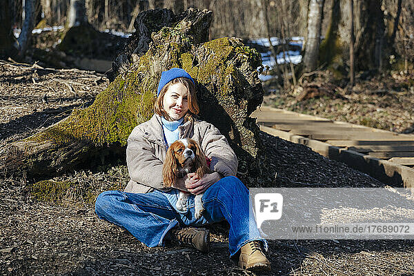 Lächelnde junge Frau mit Hund sitzt an sonnigem Tag im Wald