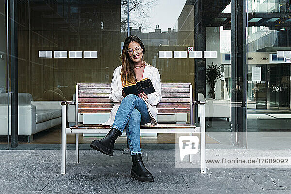 Smiling woman reading book sitting on bench in front of glass wall