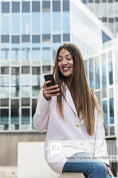 Happy woman using smart phone sitting on seat in front of building