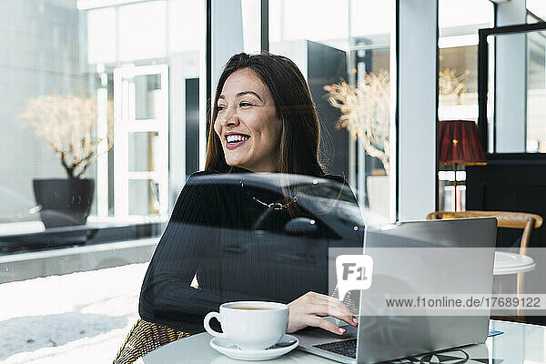 Happy businesswoman with laptop seen through glass window sitting in cafe