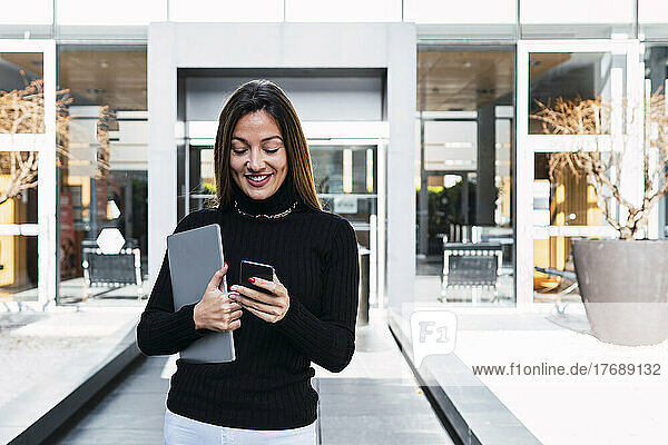 Glückliche Geschäftsfrau mit Laptop und Smartphone vor dem Café an einem sonnigen Tag