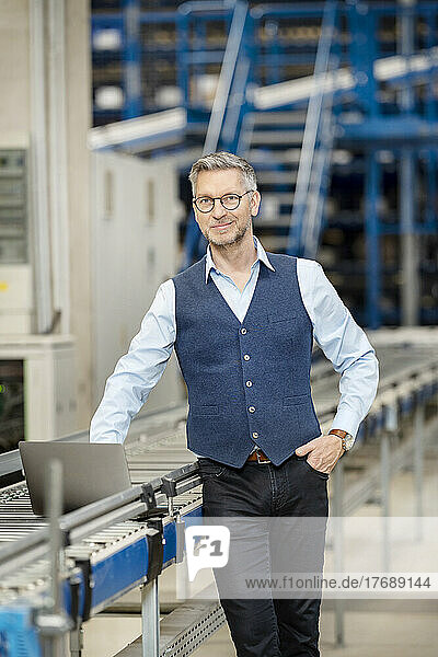 Smiling businessman with laptop on conveyor belt in warehouse