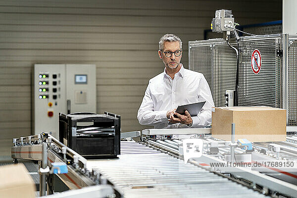 Mature businessman examining box on conveyor belt with tablet PC at factory