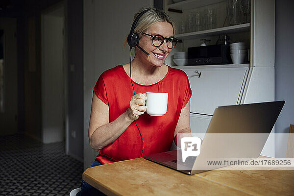 Smiling woman wearing headset using laptop holding tea cup sitting at table
