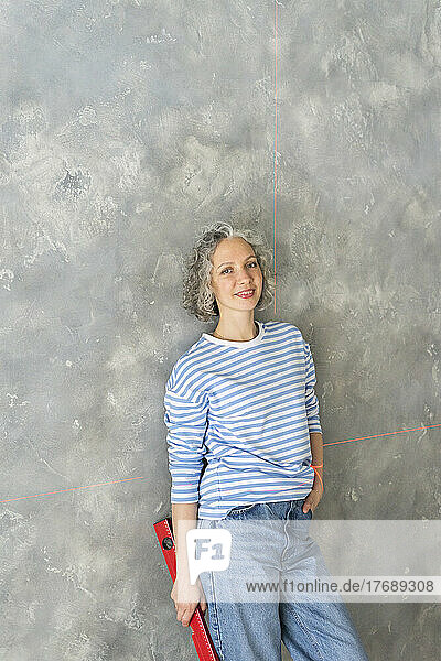 Smiling woman holding level leaning on gray wall