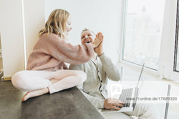 Glückliche Tochter gibt ihrem Vater  der zu Hause mit Laptop am Fenster sitzt  High-Five
