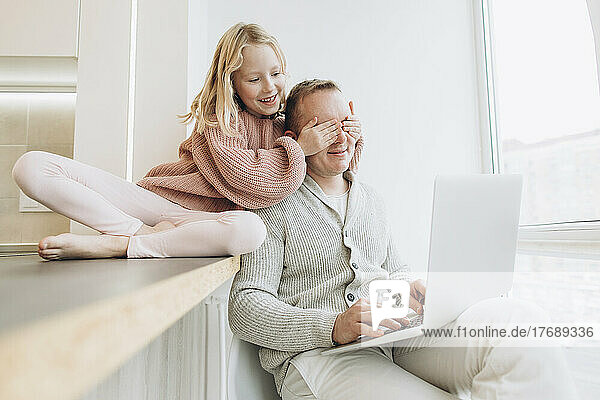 Glückliche Tochter bedeckt die Augen des Vaters  der zu Hause mit Laptop sitzt
