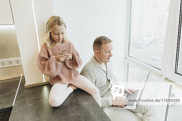 Girl using smart phone with father working on laptop in kitchen at home