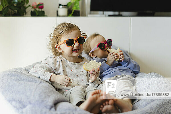 Smiling twin sisters with sunglasses having bread in living room