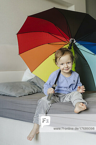 Smiling girl sitting with umbrella on bed at home