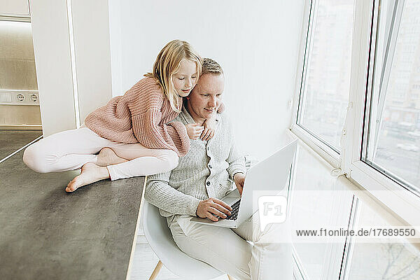 Daughter with father working on laptop sitting by window at home