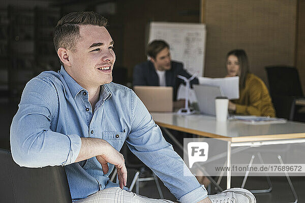 Confident businessman sitting on chair in office with colleagues in background