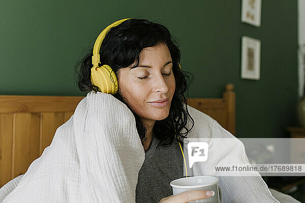 Woman with eyes closed holding tea cup listening music through headphones sitting at home