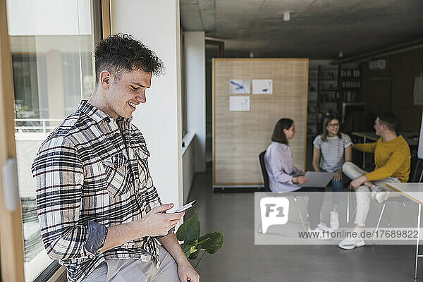 Smiling young businessman using smartphone in office with colleagues in background