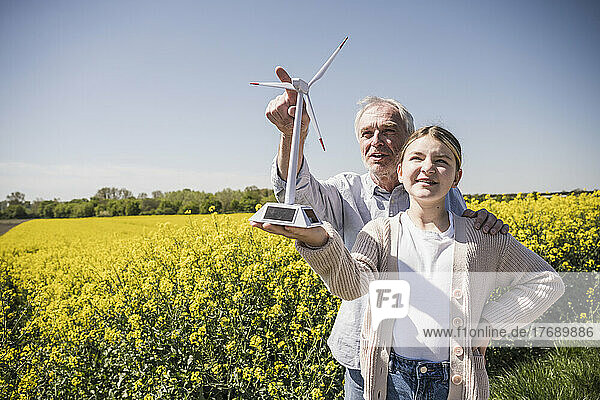 Mädchen hält Modell einer Windkraftanlage in der Hand  während Großvater auf landwirtschaftliches Feld gestikuliert