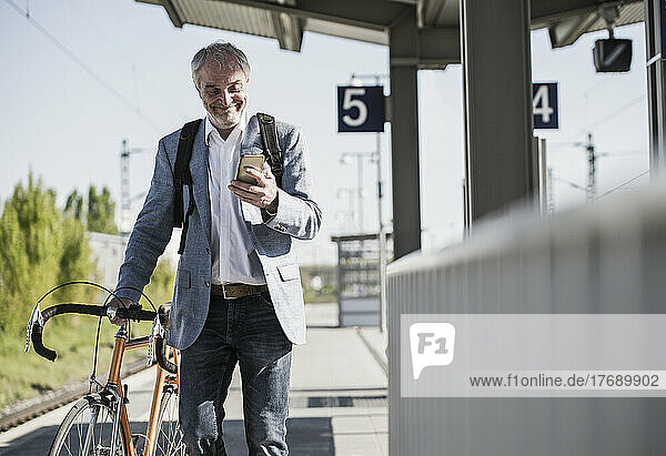 Lächelnder Geschäftsmann benutzt Smartphone und läuft mit Fahrrad auf dem Bahnsteig