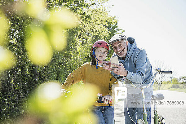 Glücklicher Großvater  der an einem sonnigen Tag sein Mobiltelefon mit seiner Enkelin teilt