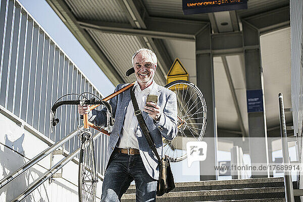 Smiling businessman carrying bicycle on shoulder holding mobile phone moving down from staircase