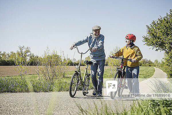 Happy grandfather and granddaughter walking with bicycle on road