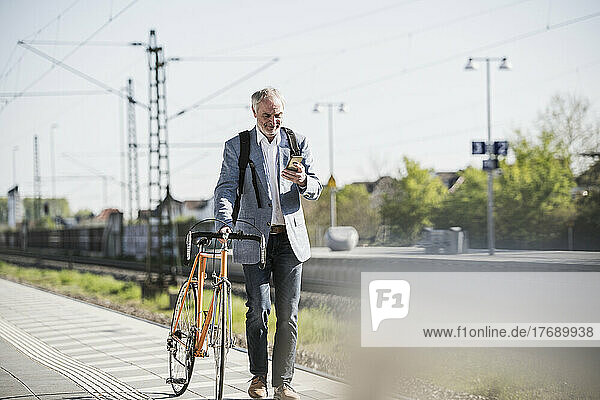 Businessman using smart phone walking with bicycle on railroad station platform
