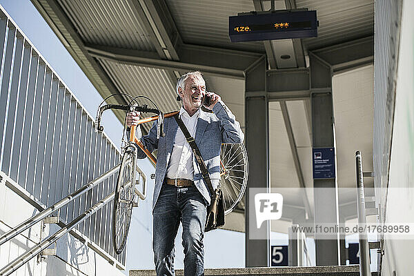 Lächelnder Geschäftsmann trägt Fahrrad auf der Schulter und spricht am Bahnhof per Smartphone