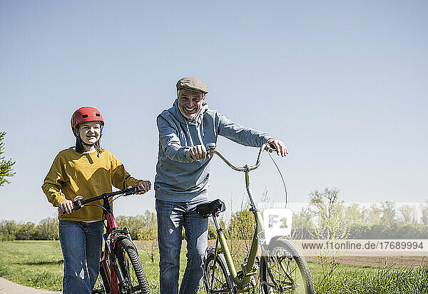 Happy grandfather and granddaughter walking with bicycle on sunny day