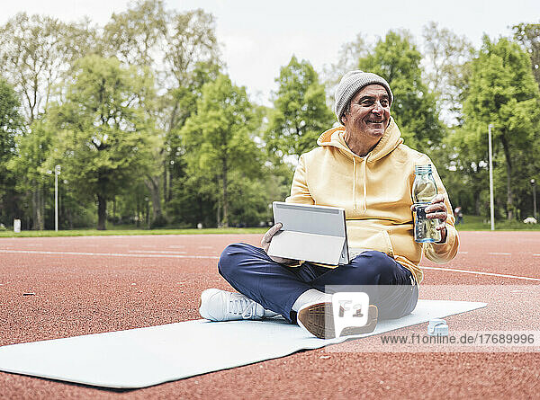 Smiling senior man with tablet PC and water bottle sitting on exercise mat