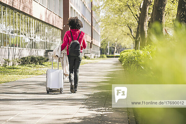 Businesswoman with backpack and wheeled luggage walking on footpath