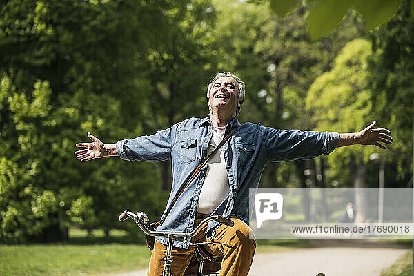 Carefree senior man with arms outstretched riding bicycle at park