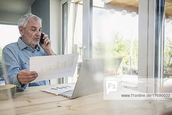 Senior businessman with documents talking on mobile phone at home