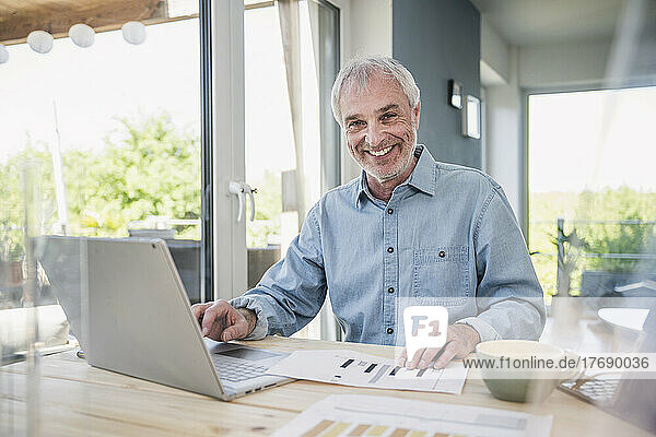 Happy senior businessman with laptop and graph sitting at table
